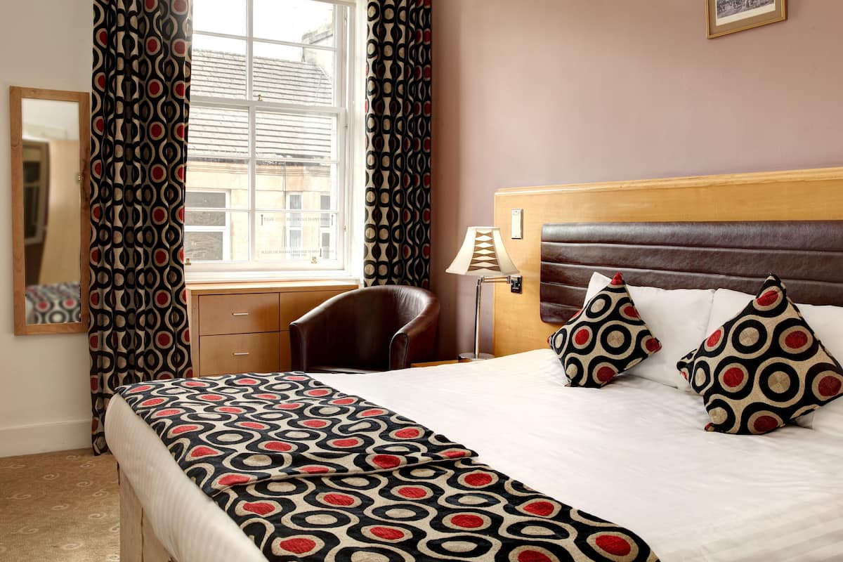 Ein Hotelzimmer mit Bett, Sessel, Schreibtisch, Lampe und einem fenster mit Vorhängen.