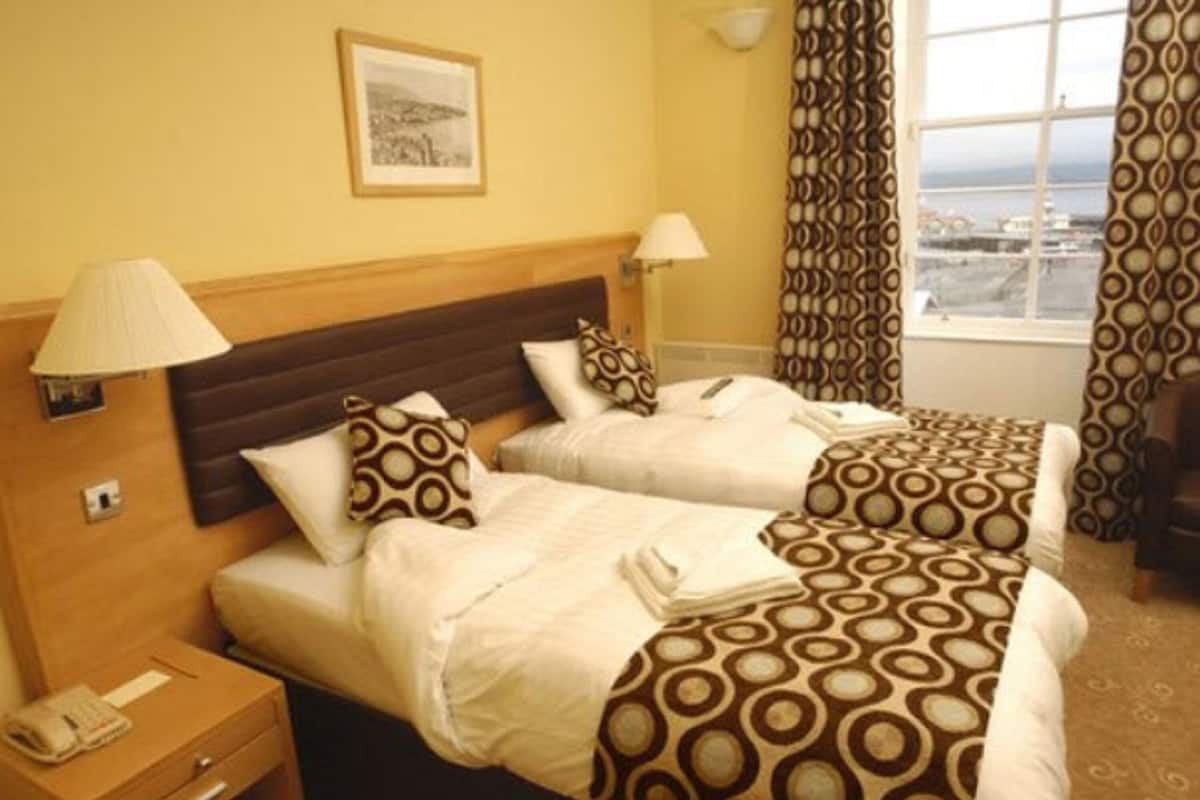 Ein Hotelzimmer mit zwei Betten, einem Holzkopfende, einem Nachttisch mit Lampe und einem Fenster mit Vorhängen.