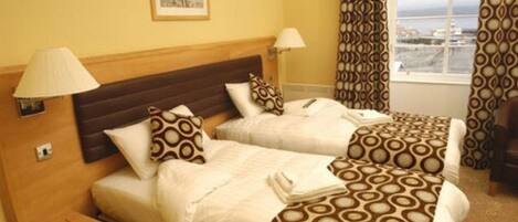 A hotel room with two beds, a wooden headboard, a bedside table with a lamp, and a window with curtains.