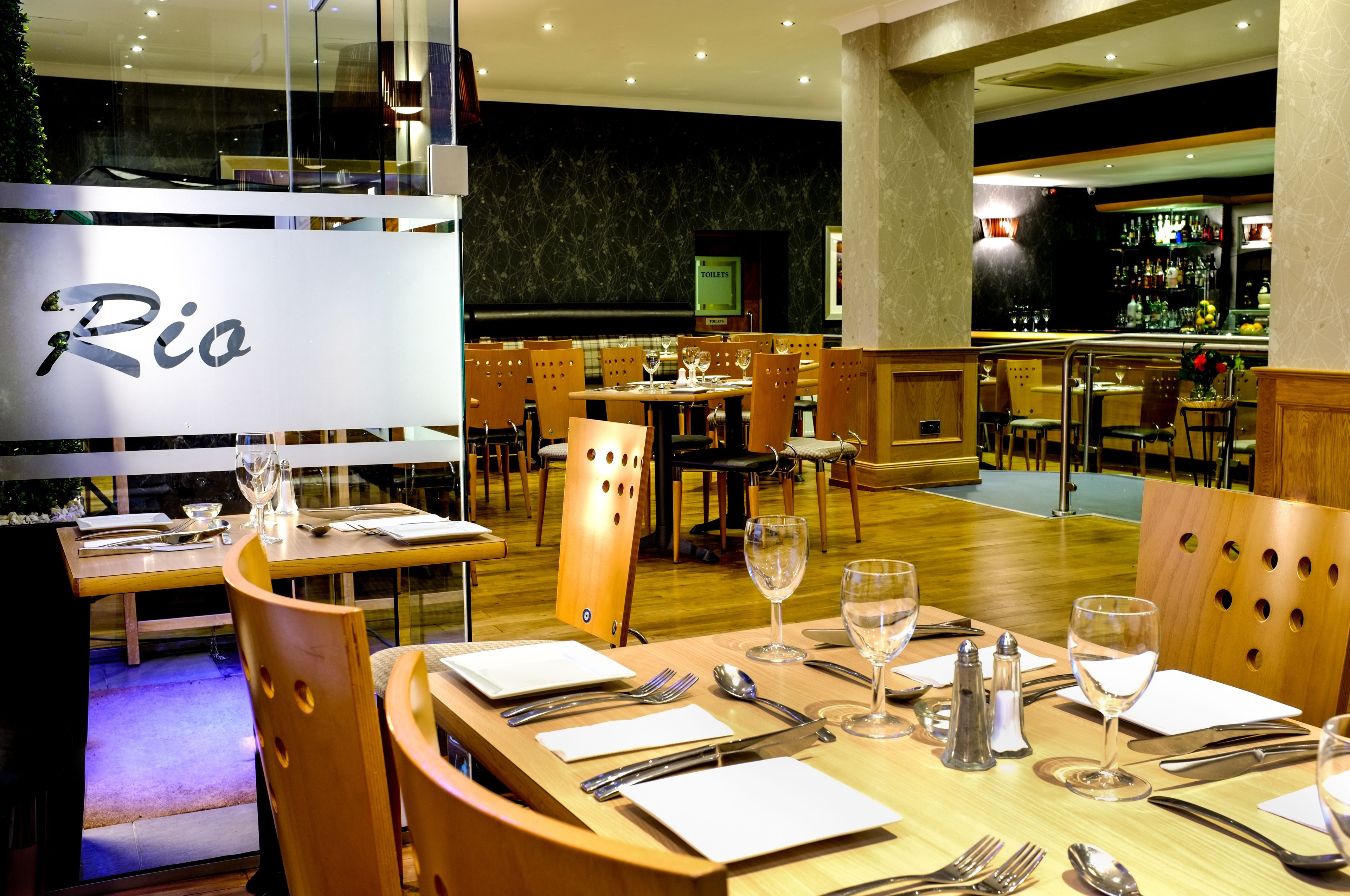 Ein Restaurant mit mehreren Essplätzen, jeweils gedeckt mit Besteck und Weingläsern, und im Hintergrund ist eine Bar zu sehen.