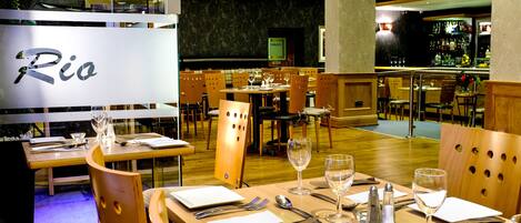 A restaurant with multiple dining tables, each set with cutlery and wine glasses, and a bar area visible in the background.