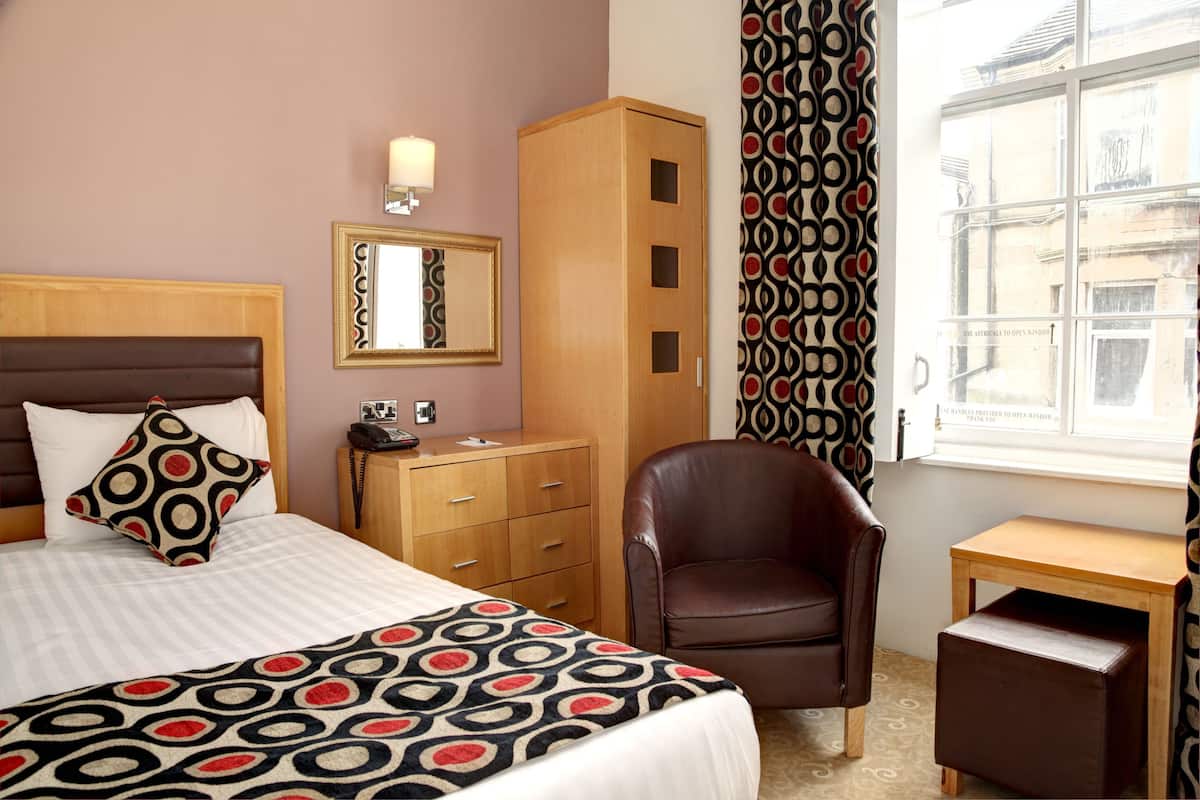 Ein Hotelzimmer mit einem Bett, einem Holzschrank, einem Ledersessel, einem kleinen Holztisch und einem Fenster mit gemusterten Vorhängen.