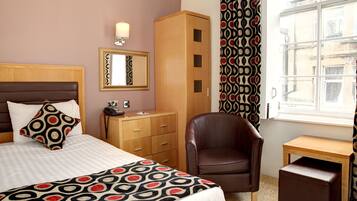 Una habitación de hotel con cama, un armario de madera, un sillón de cuero, una mesita de madera y una ventana con cortinas estampadas.