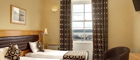 A hotel room with two beds, a chair, a desk, a lamp, and a window with curtains.