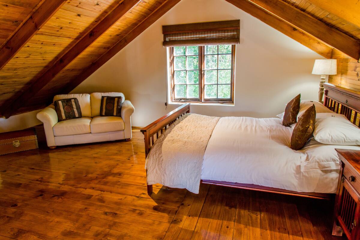 A bedroom with a wooden ceiling, a bed with white linens, a wooden nightstand, a wooden dresser, and a window with a view of greenery.