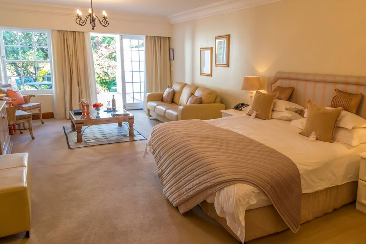 A spacious bedroom with a large bed, a sitting area with a sofa and armchairs, a dining table, and a view of the outdoors through a large window.