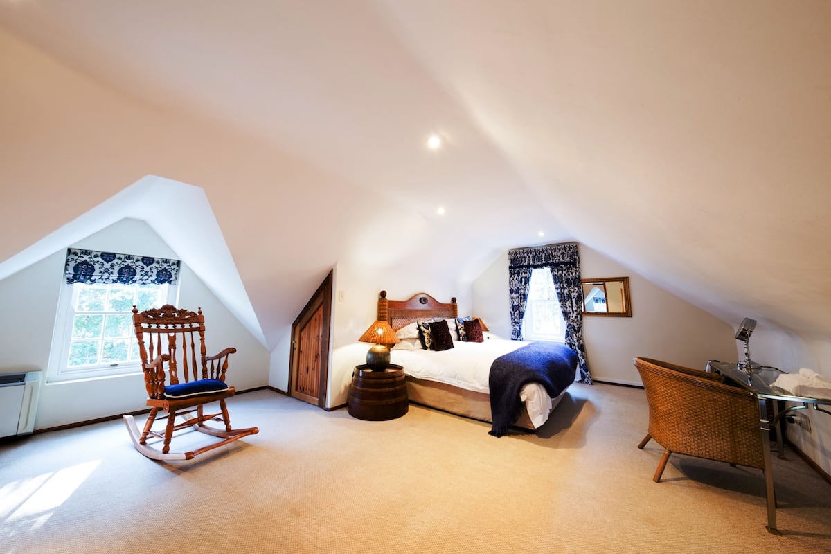A bedroom with a sloped ceiling, a bed with a blue blanket, a rocking chair, a small table with a lamp, and a wicker chair.