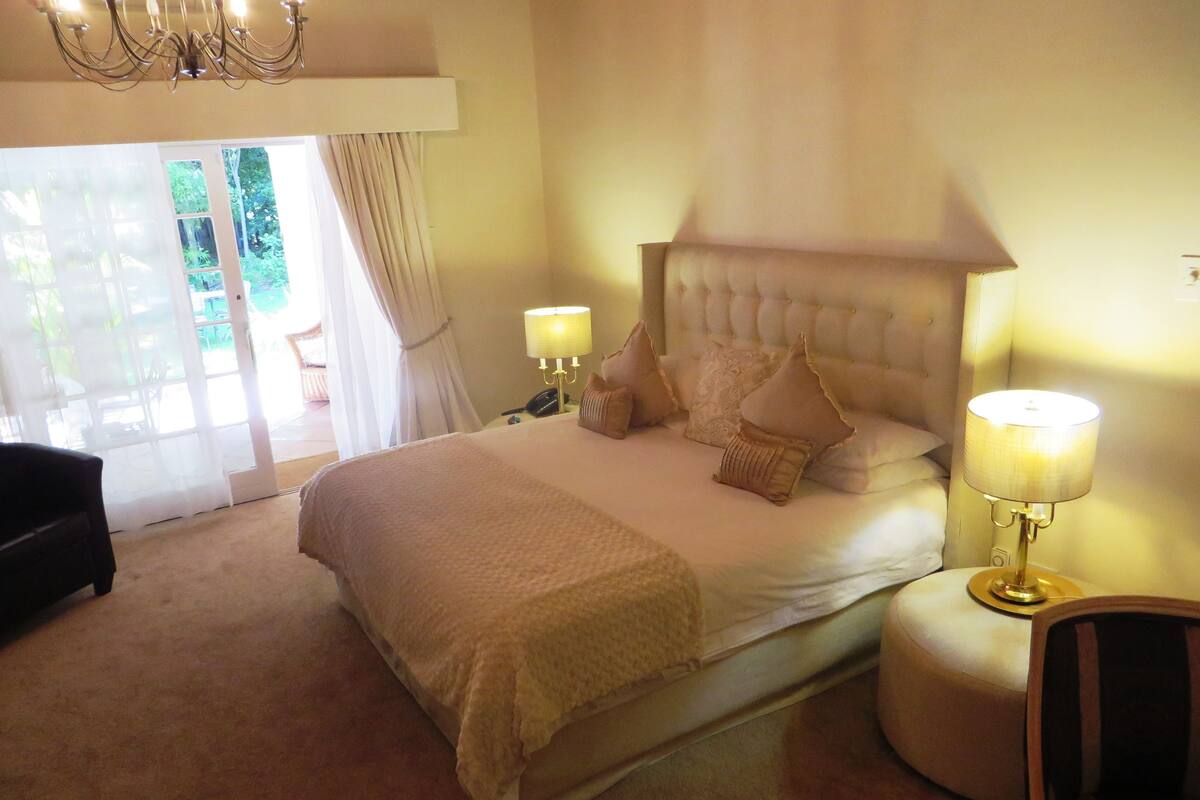 A bedroom with a large bed, two bedside lamps, a chandelier, and a view through a glass door.