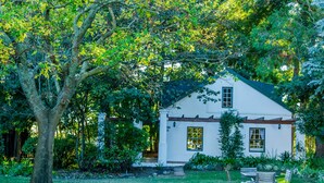 Una casa blanca con techo verde, rodeada de abundante vegetación y un gran árbol.