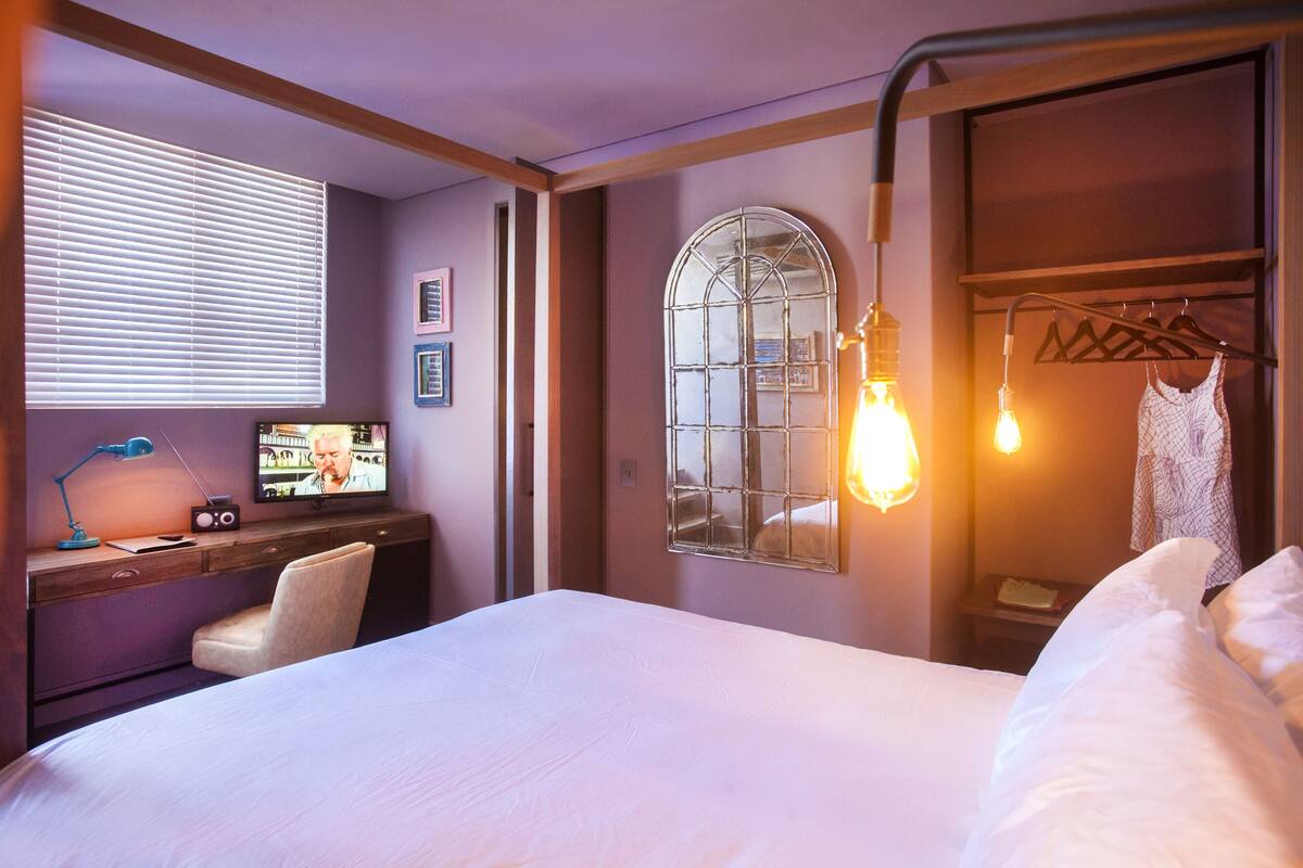 A hotel room with a bed, a desk with a computer, a chair, a TV, and a window with blinds.