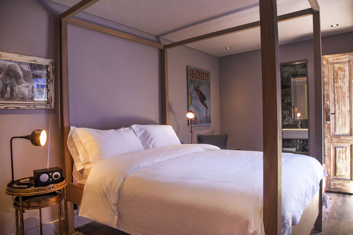 A bedroom with a four-poster bed, a bedside table with a lamp, a radio, and a poster on the wall.