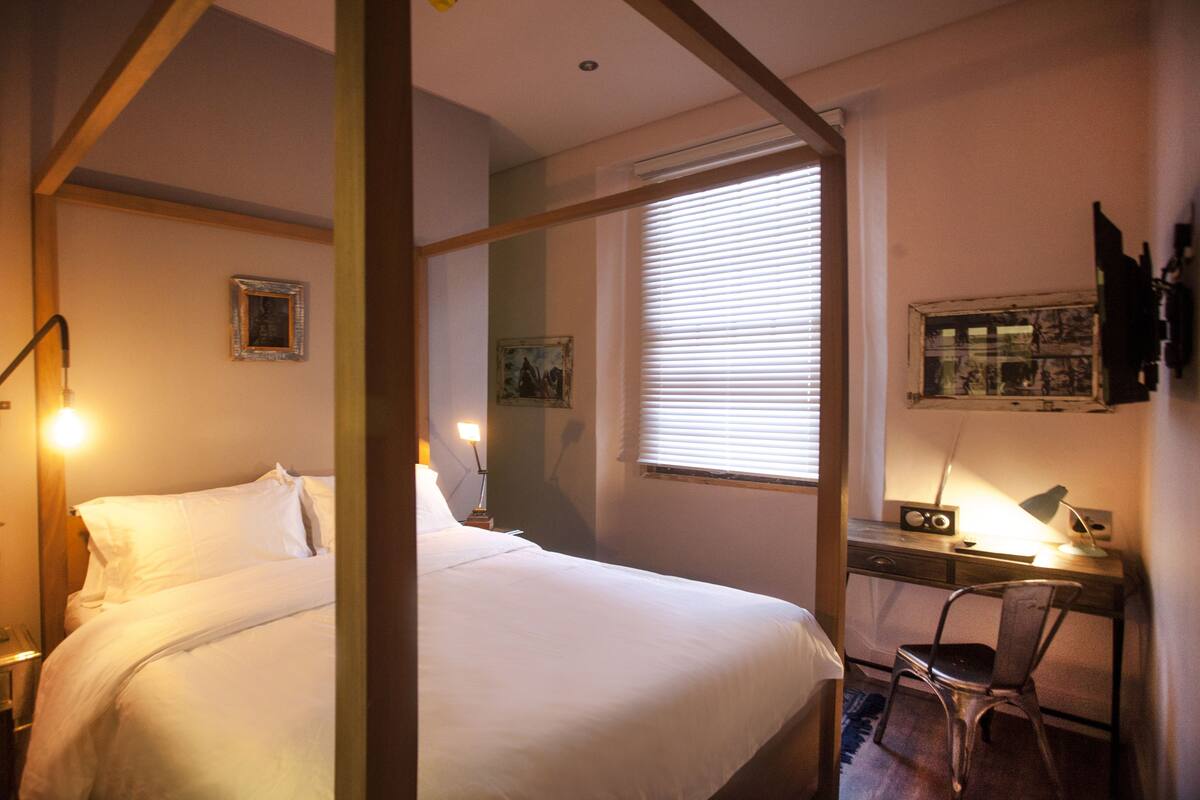 A hotel room with a bed, a desk, and a chair. There are two lamps on the desk and a picture frame on the wall.