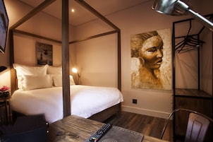 Habitación de hotel con cama con dosel, un escritorio con silla, televisor montado y un cuadro abstracto grande en la pared.