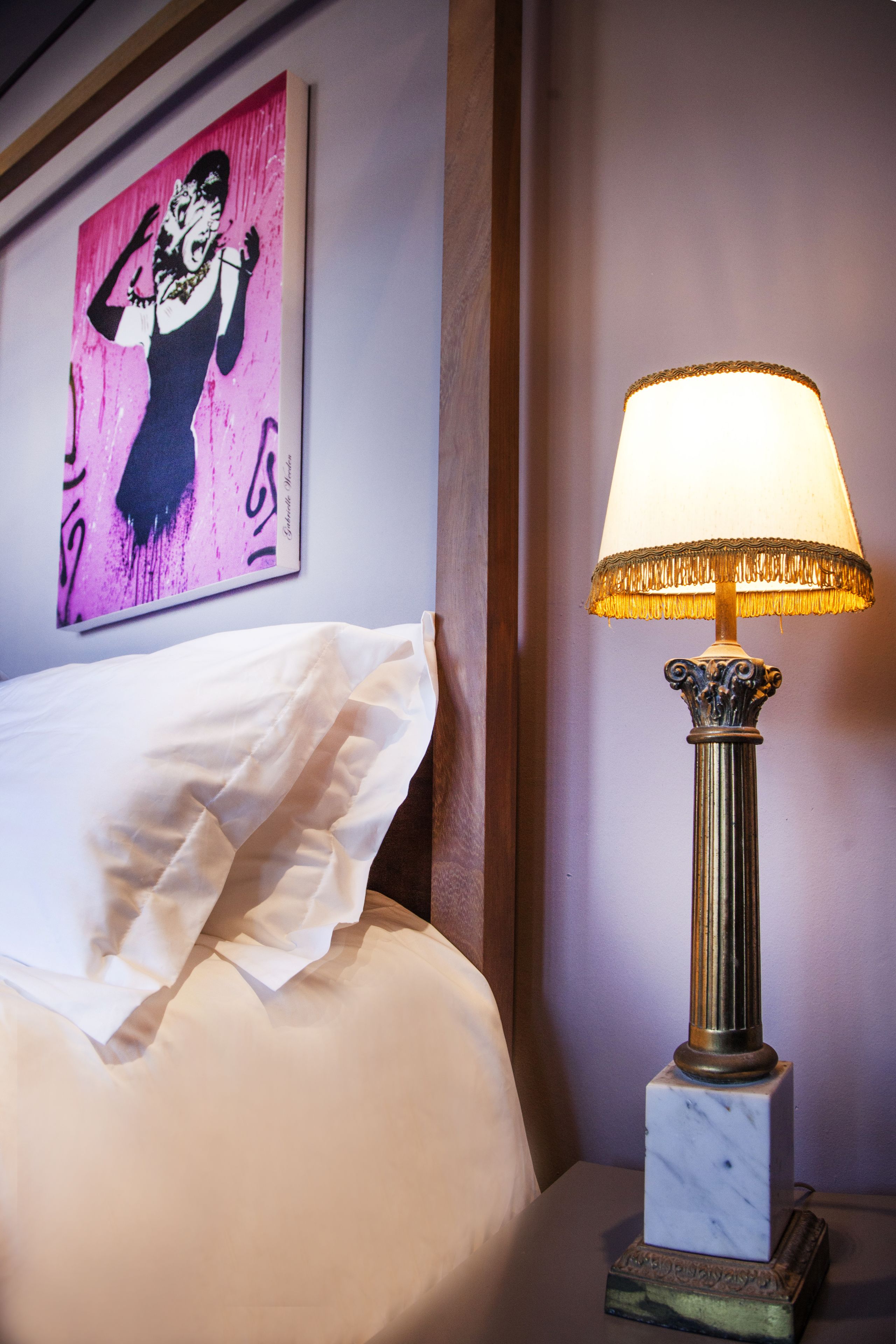 A neatly made bed with white linens, a decorative lamp with a gold base and fringed lampshade, and a pink artwork on the wall.