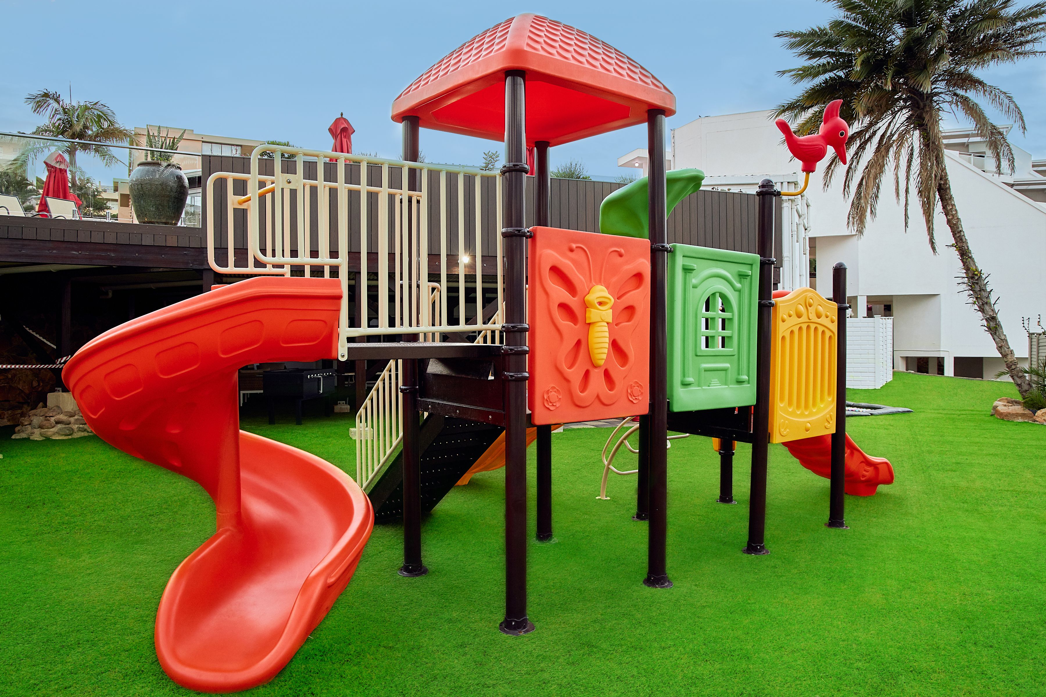 A playground with a red slide, a climbing structure, and a small red bird figure.