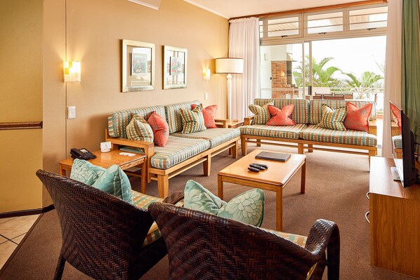 A hotel room with a sofa set, a dining table, wicker chairs, a TV, and a view of the outdoors.