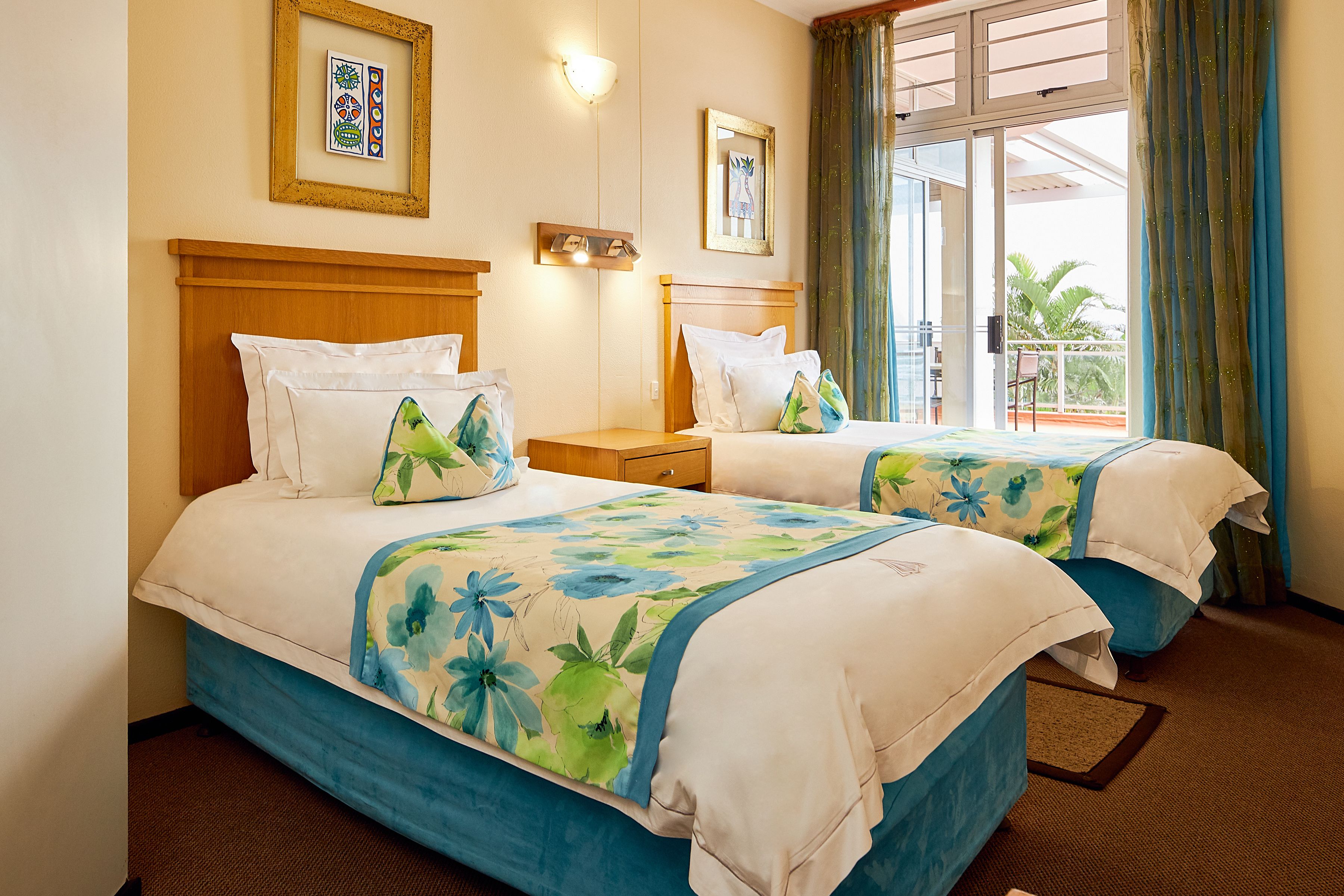 A hotel room with two beds, a wooden headboard, a nightstand, and a balcony with a view of palm trees.