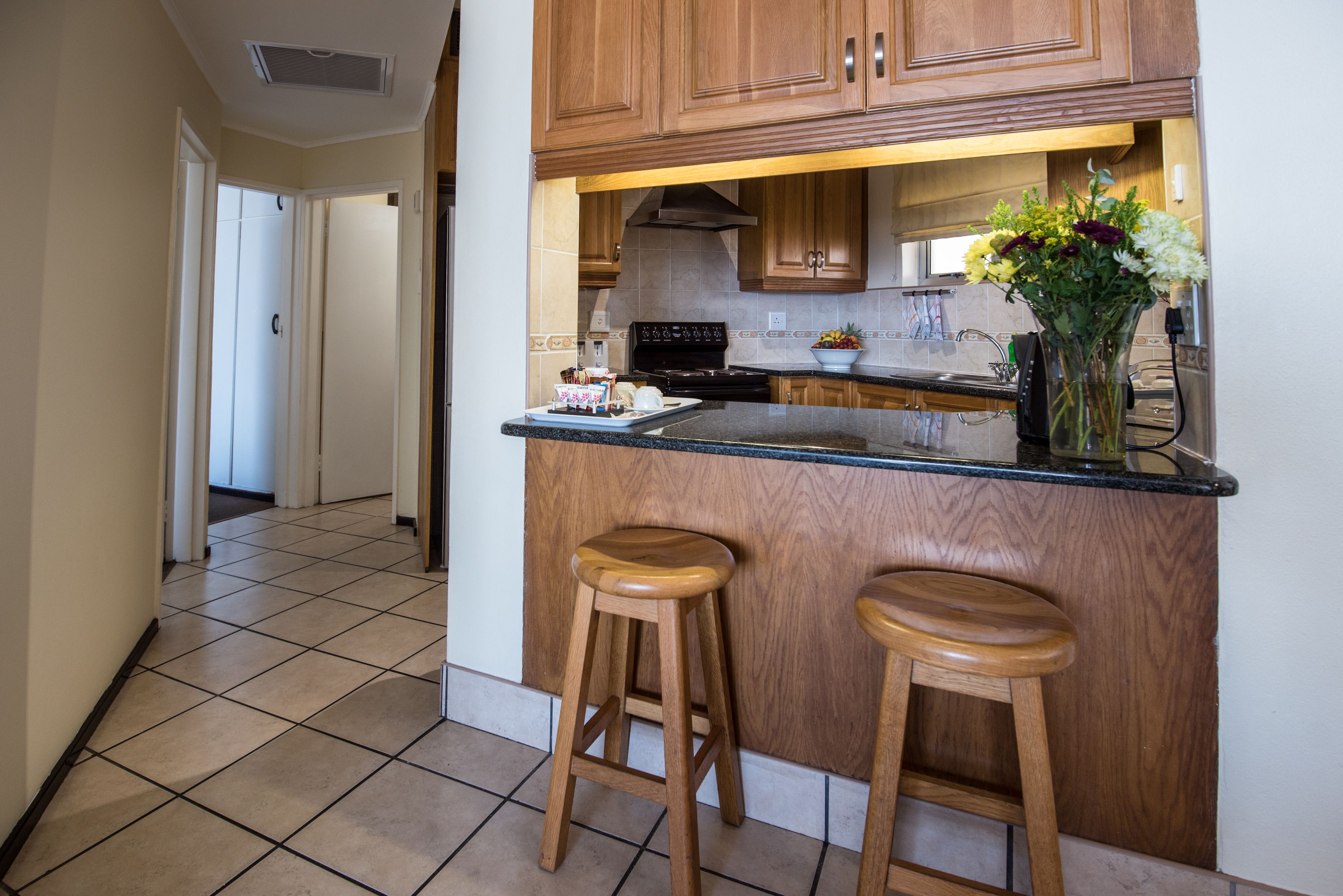 A kitchen with wooden cabinets, a black countertop, two wooden stools, and a vase with flowers.