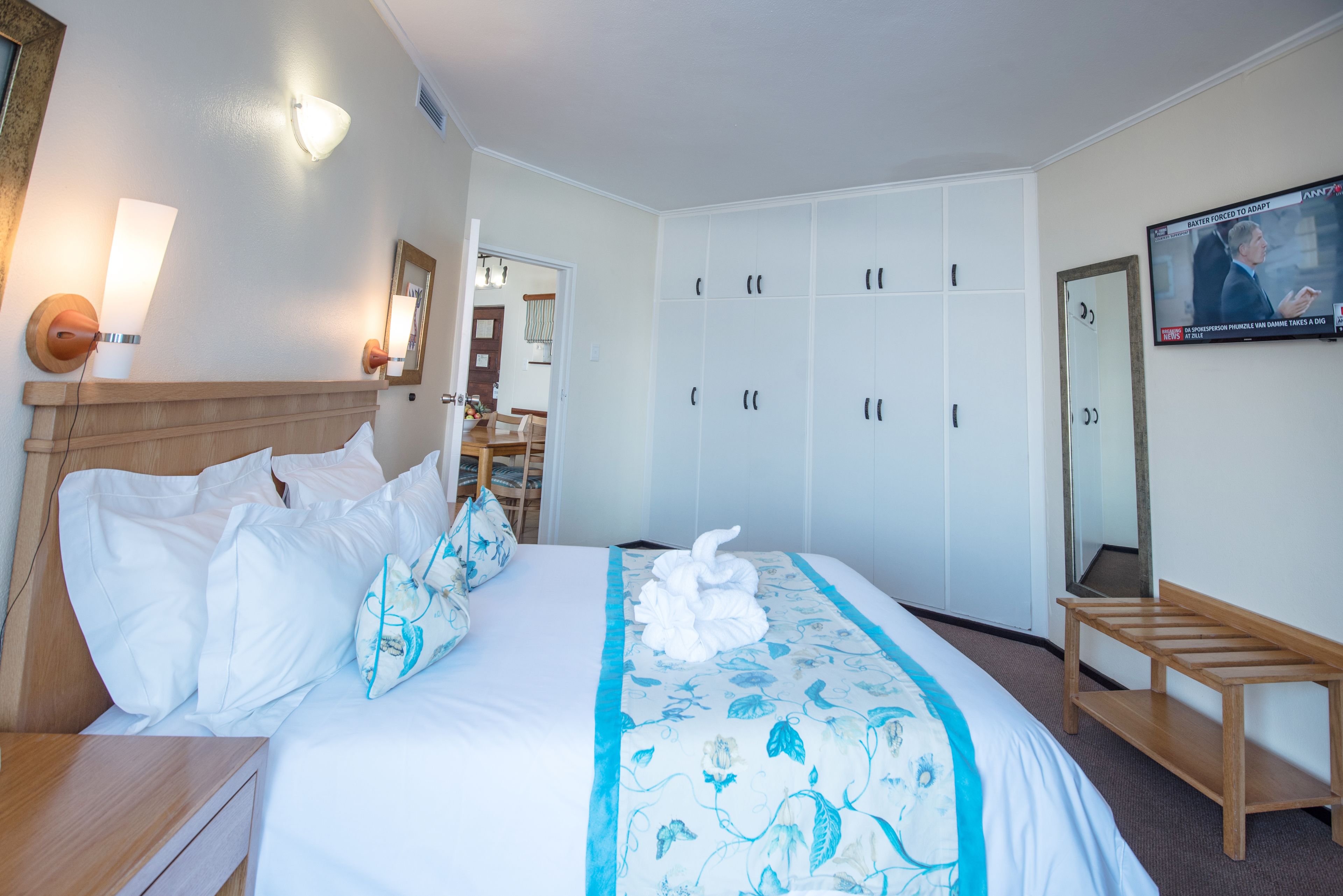 A hotel room with a bed, a wooden nightstand, a mirror, a wardrobe, and a television on the wall.