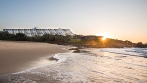 Un hôtel situé en bord de mer, avec un bâtiment aux lignes courbes, une plage de sable et des vagues qui se brisent sur le rivage.