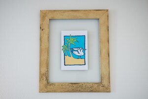 Una obra de arte enmarco que muestra una playa, palmeras y un ave.