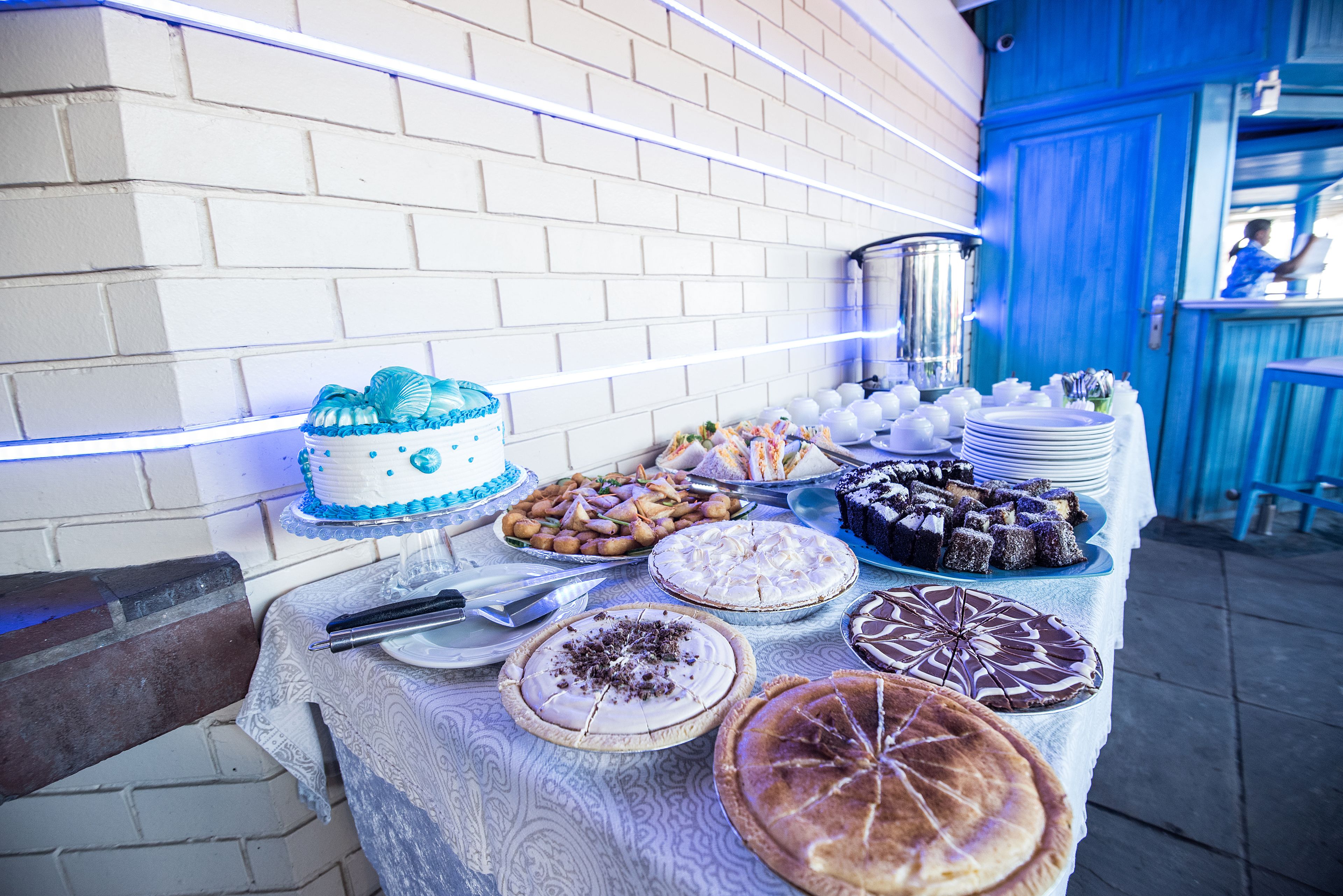Una mesa con diversos postres y un pastel, frente a una pared de ladrillo con iluminación azul.