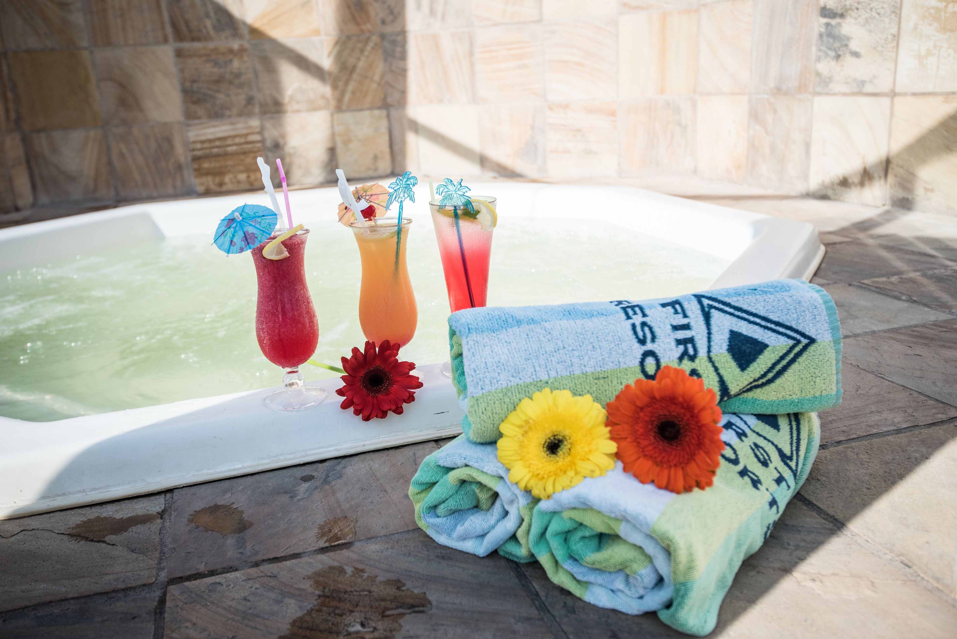 A bathtub with two drinks, a towel with flowers, and a red flower on a stone floor.