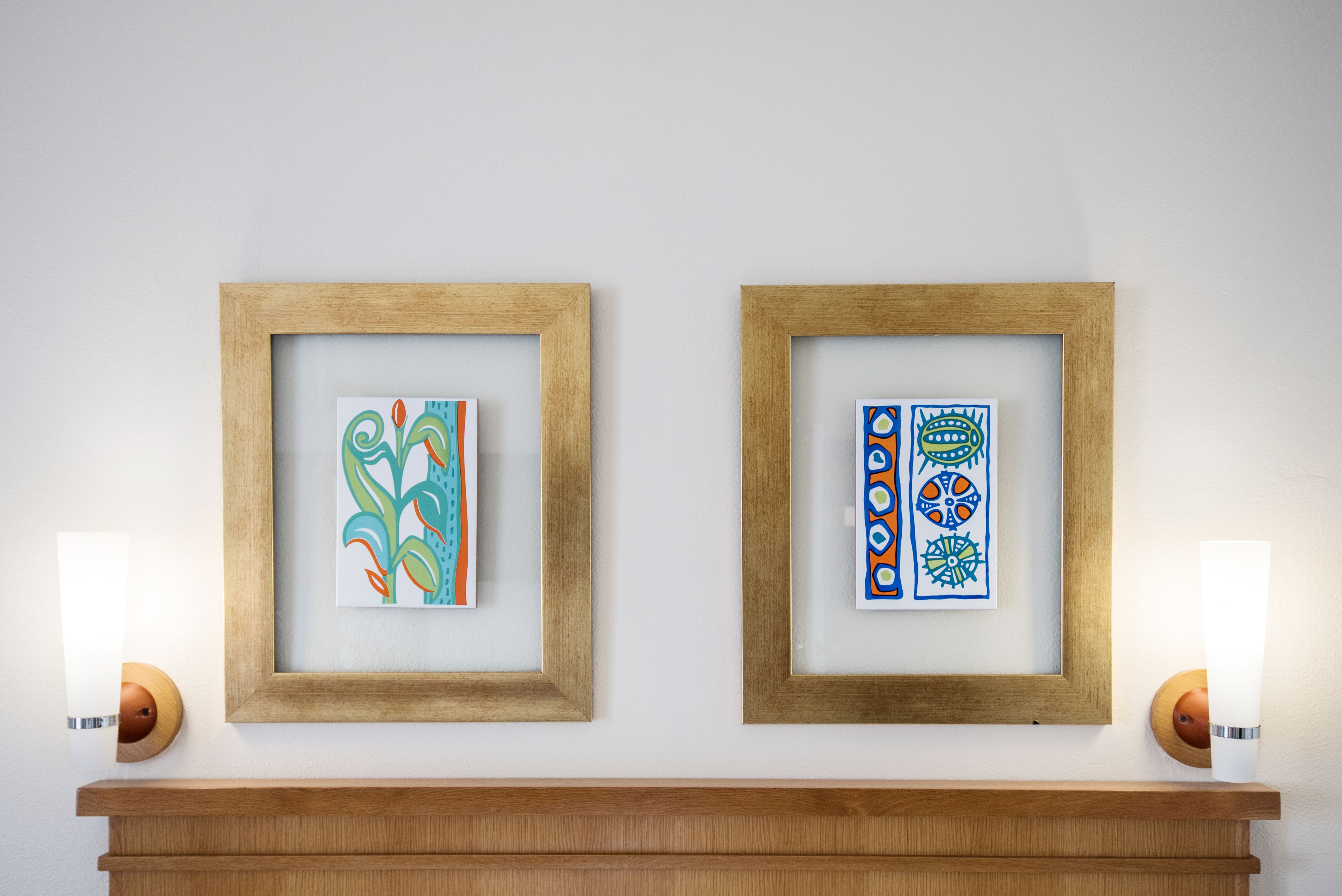 Two framed artworks on a wall above a wooden shelf.