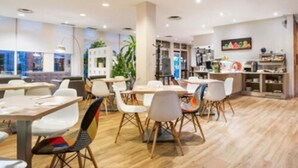 A modern dining area with white tables and chairs, a kitchen in the background, and a large window with a view. - Hotel Majadahonda (Majadahonda)