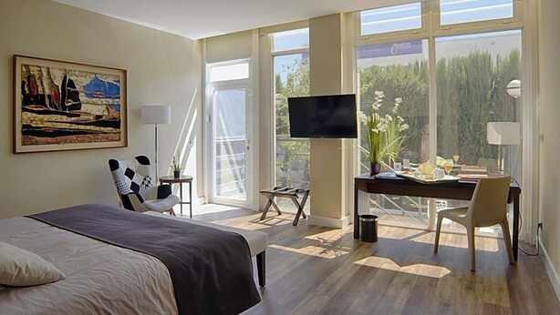 A hotel room with a bed, a desk with a TV, a chair, and a window with a view of trees. - Hotel Majadahonda (Majadahonda)