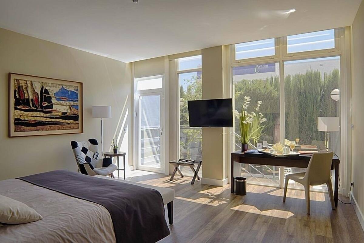 A hotel room with a bed, a desk with a TV, a chair, and a window with a view of trees.