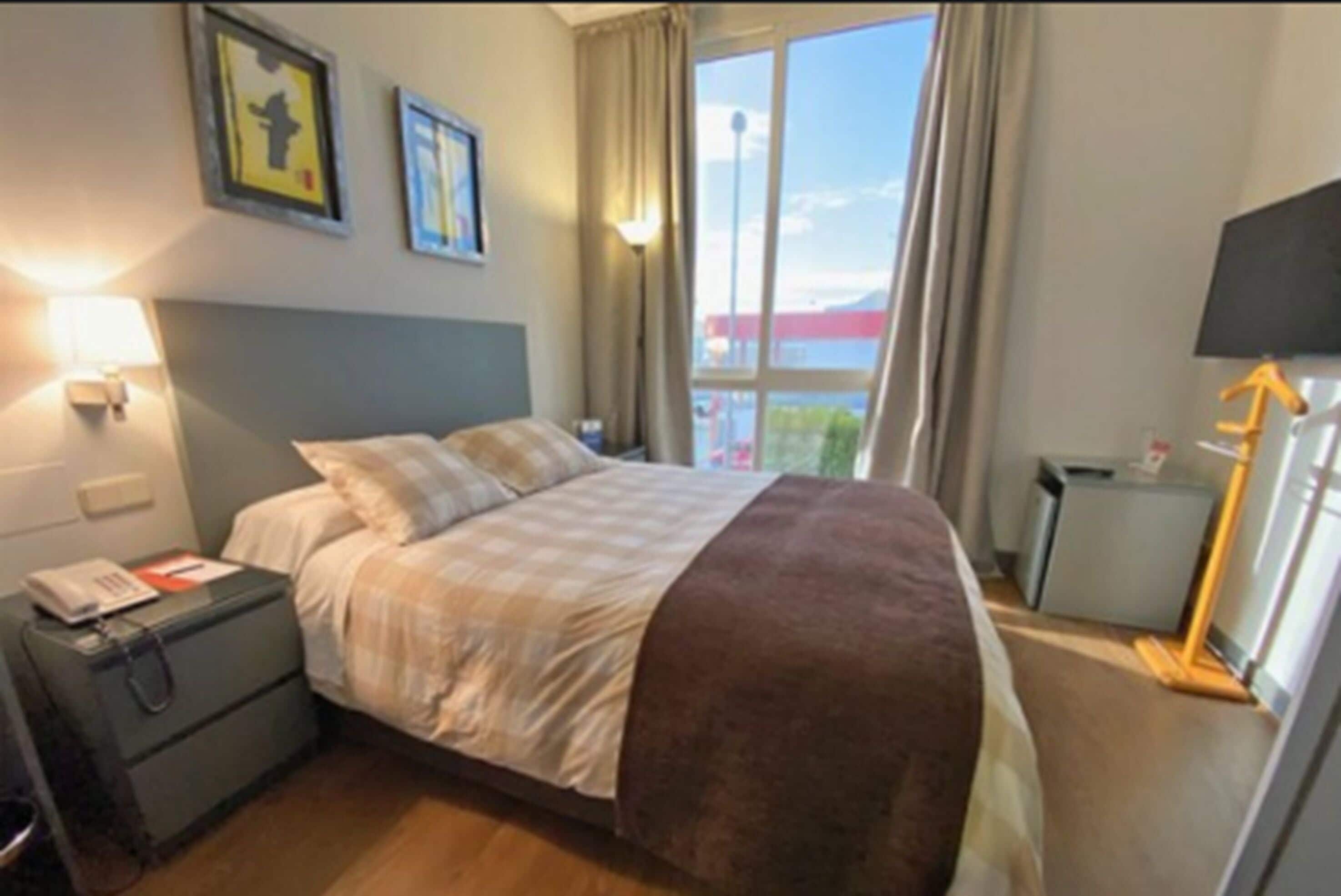 Habitación de hotel con cama, mesita de noche con lámpara, pequeño escritorio con teléfono y ventana con vista al exterior.