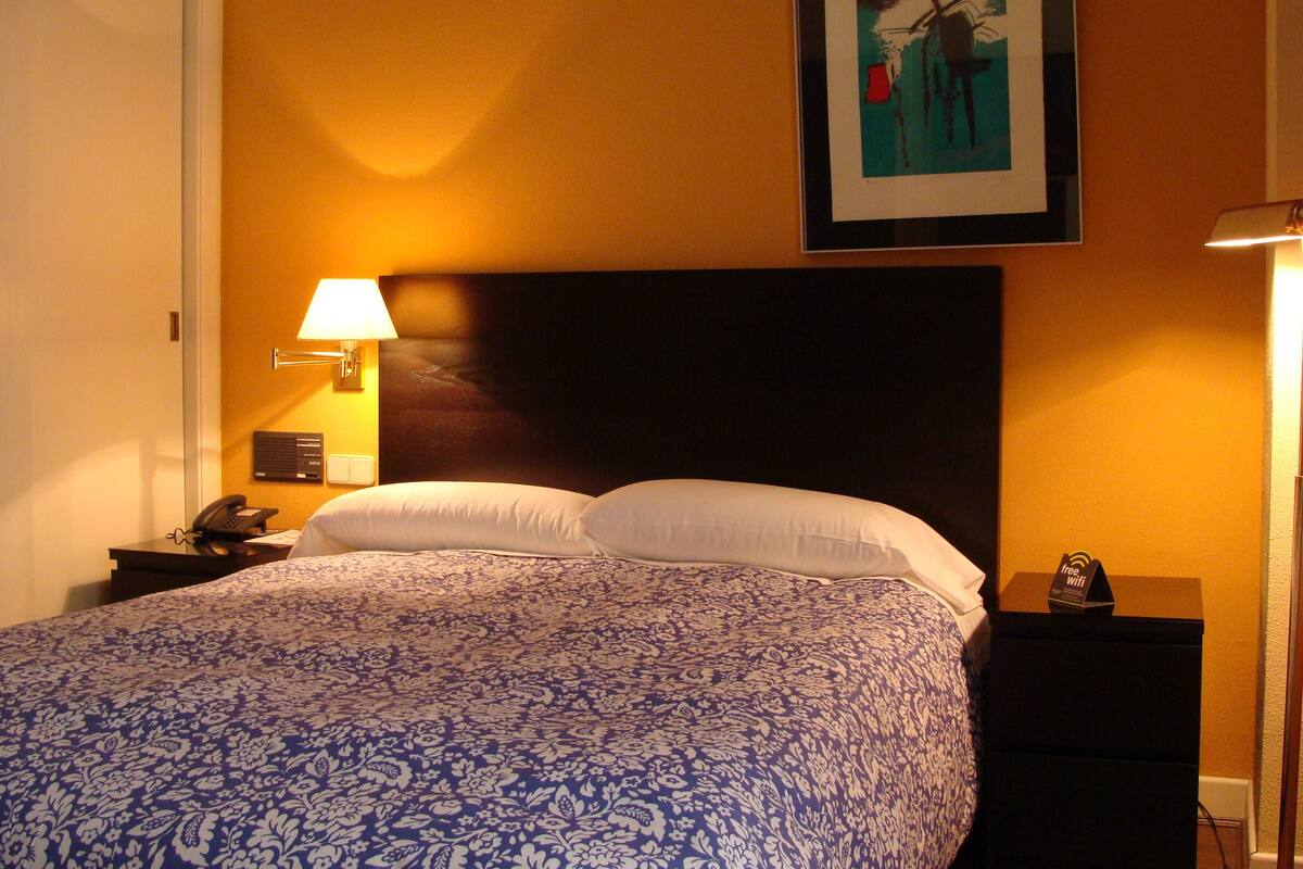 A hotel room with a large bed, a nightstand with a lamp, a phone, and a framed picture on the wall.