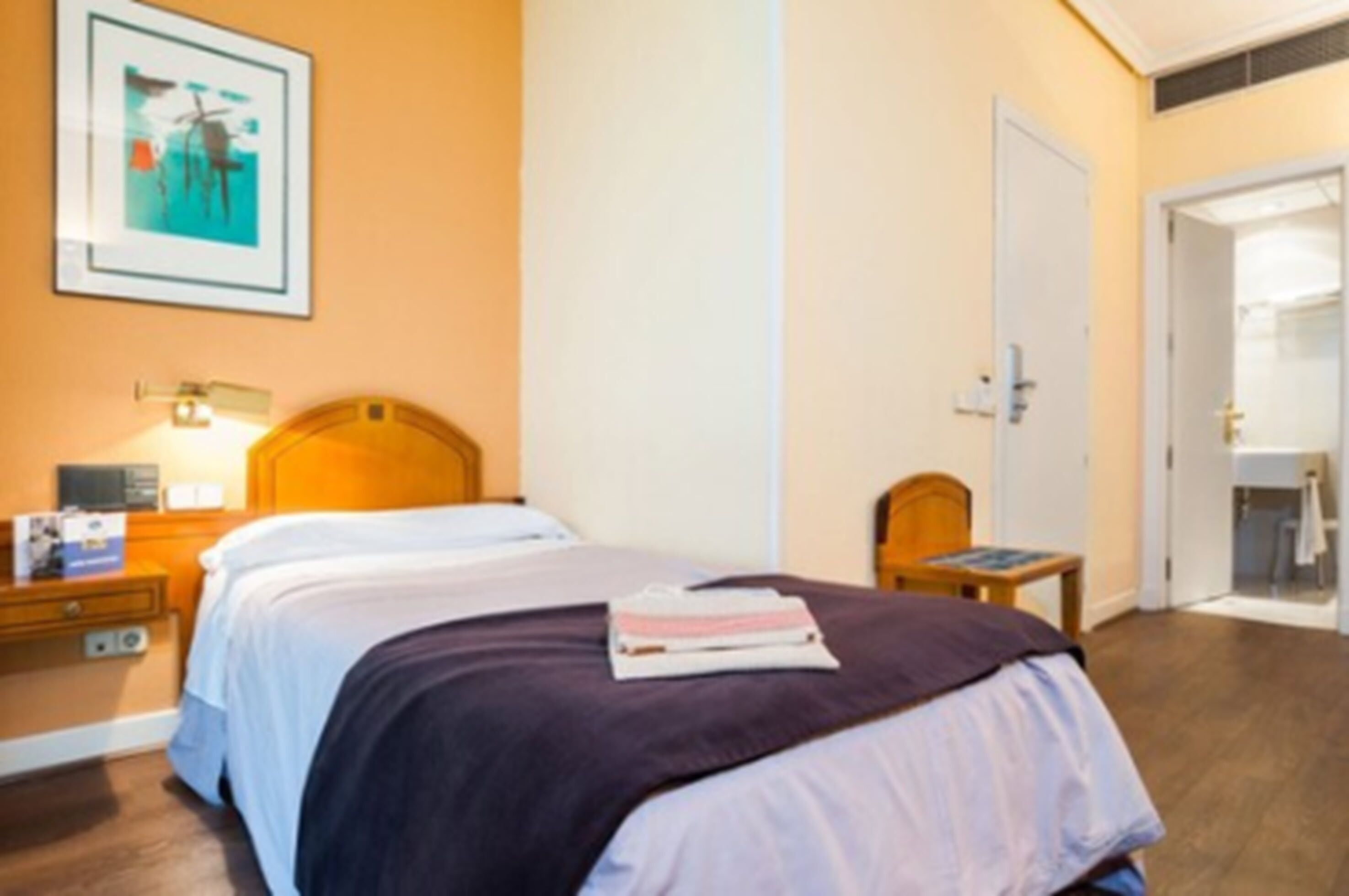 A hotel room with a bed, a nightstand, a wall-mounted lamp, a door, and a framed picture on the wall.
