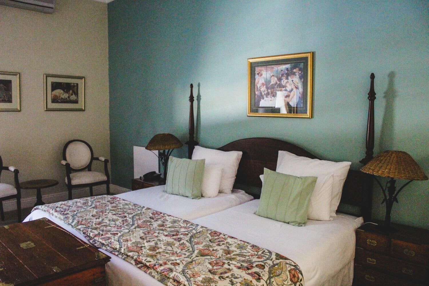 Una cama doble con cubrecama floral, dos mesitas de noche con lámparas, una silla y dos cuadros enmarcados en la pared.