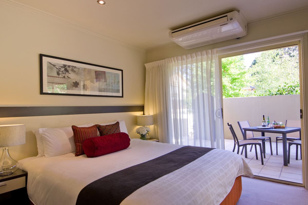 A hotel room with a large bed, a nightstand with a lamp, a framed picture on the wall, and a sliding glass door leading to an outdoor area with a table and chairs.