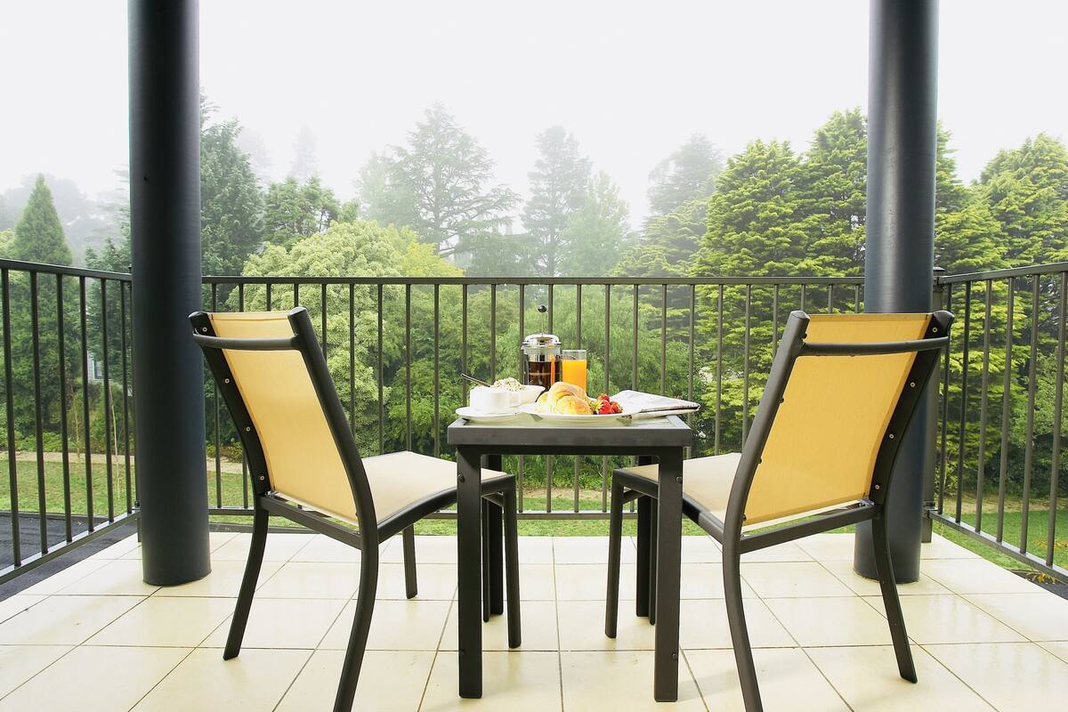 A balcony with two chairs and a table, overlooking a garden with trees and a fence.