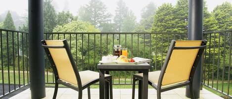 A balcony with two chairs and a table, overlooking a garden with trees and a fence.