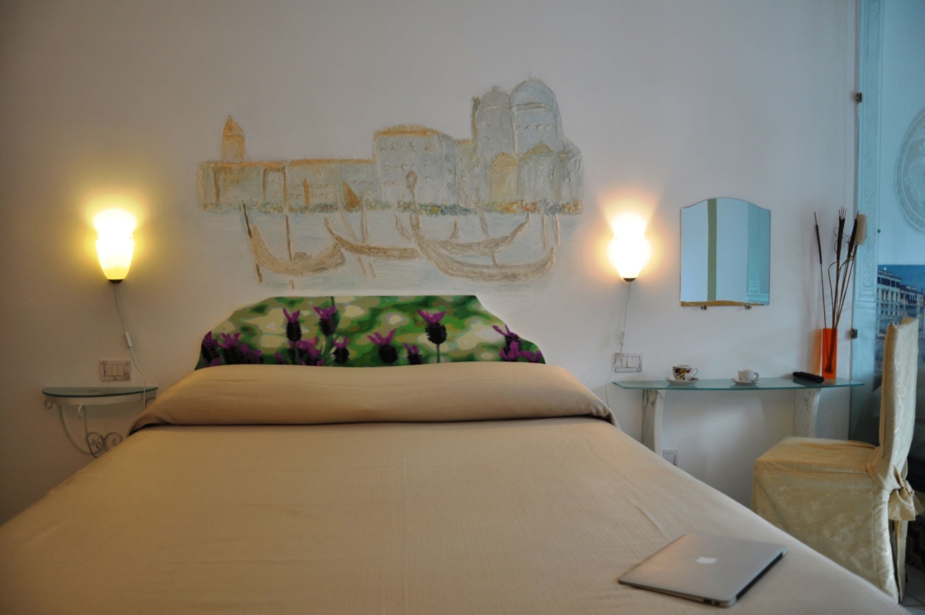 Habitación de hotel con cama, dos lámparas de noche, una mesita con taza y platillo, y una silla.