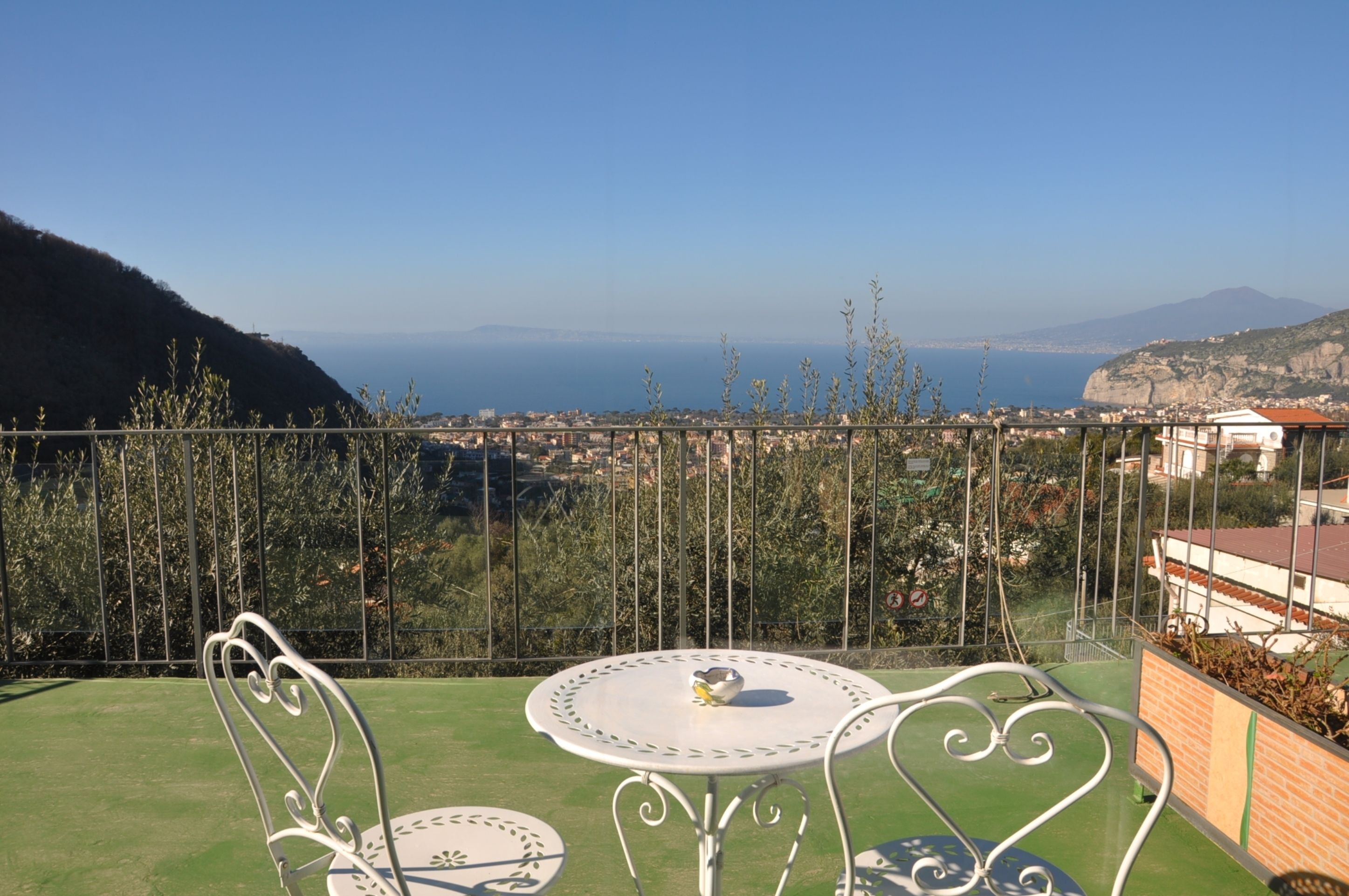 Un balcón con una mesa y sillas de metal, con vistas a una ciudad costera y montañas al fondo.