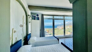 Een hotelkamer met twee bedden, een groot raam met uitzicht op zee en een bergachtig landschap, en een balkon met een hekwerk.