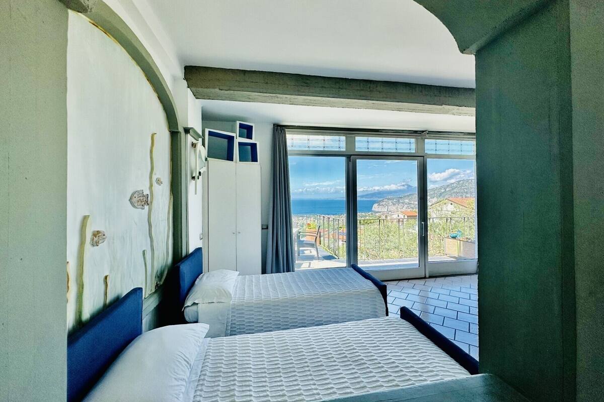 A hotel room with two beds, a large window with a view of the sea and a mountainous landscape, and a balcony with a railing.
