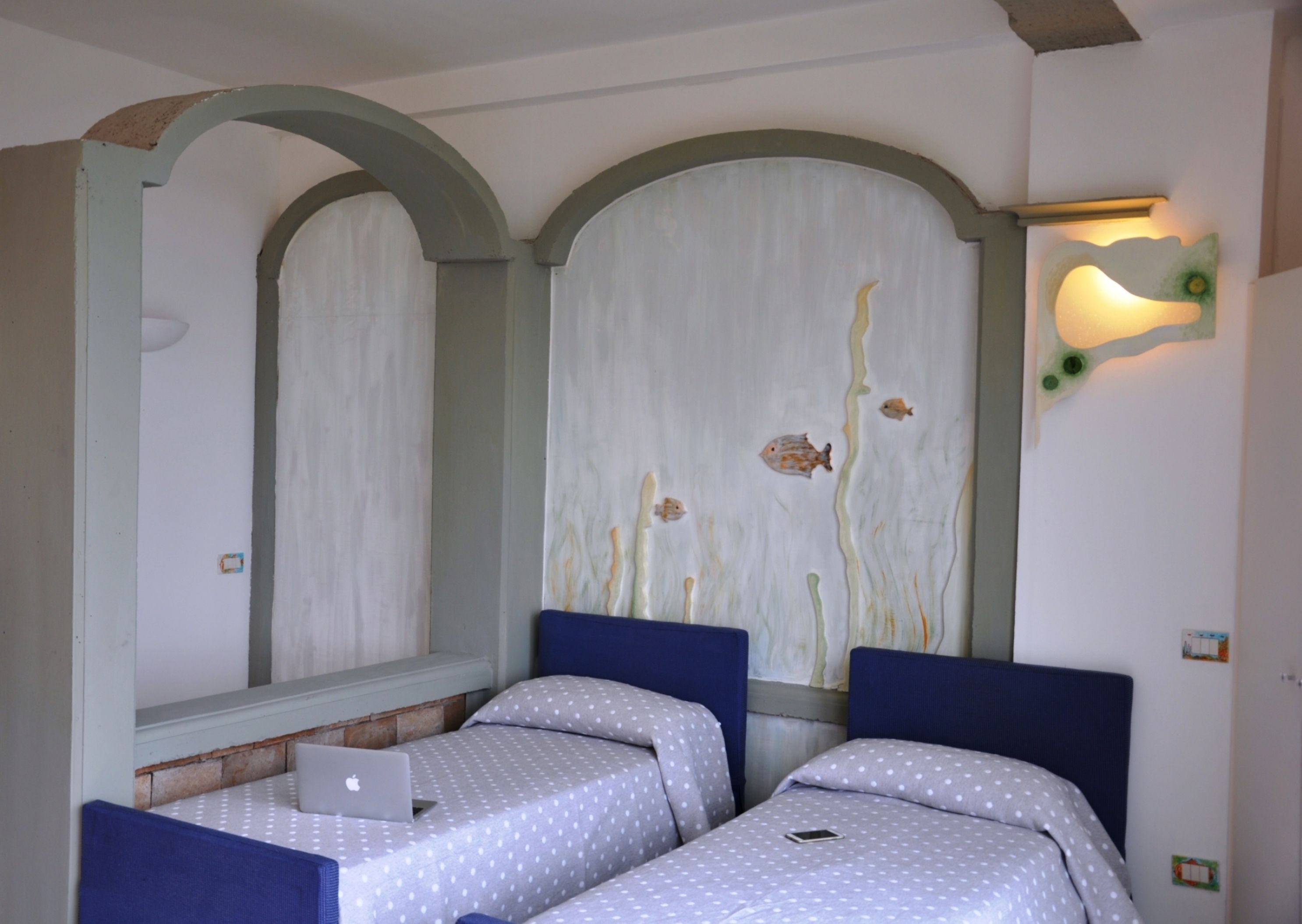 A room with two beds, each with a blue headboard and white bedding with polka dots. There is a laptop on one bed and a mobile phone on the other. The walls feature a large mural with fish and abstract designs.