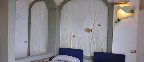 A room with two beds, each with a blue headboard and white bedding with polka dots. There is a laptop on one bed and a mobile phone on the other. The walls feature a large mural with fish and abstract designs.