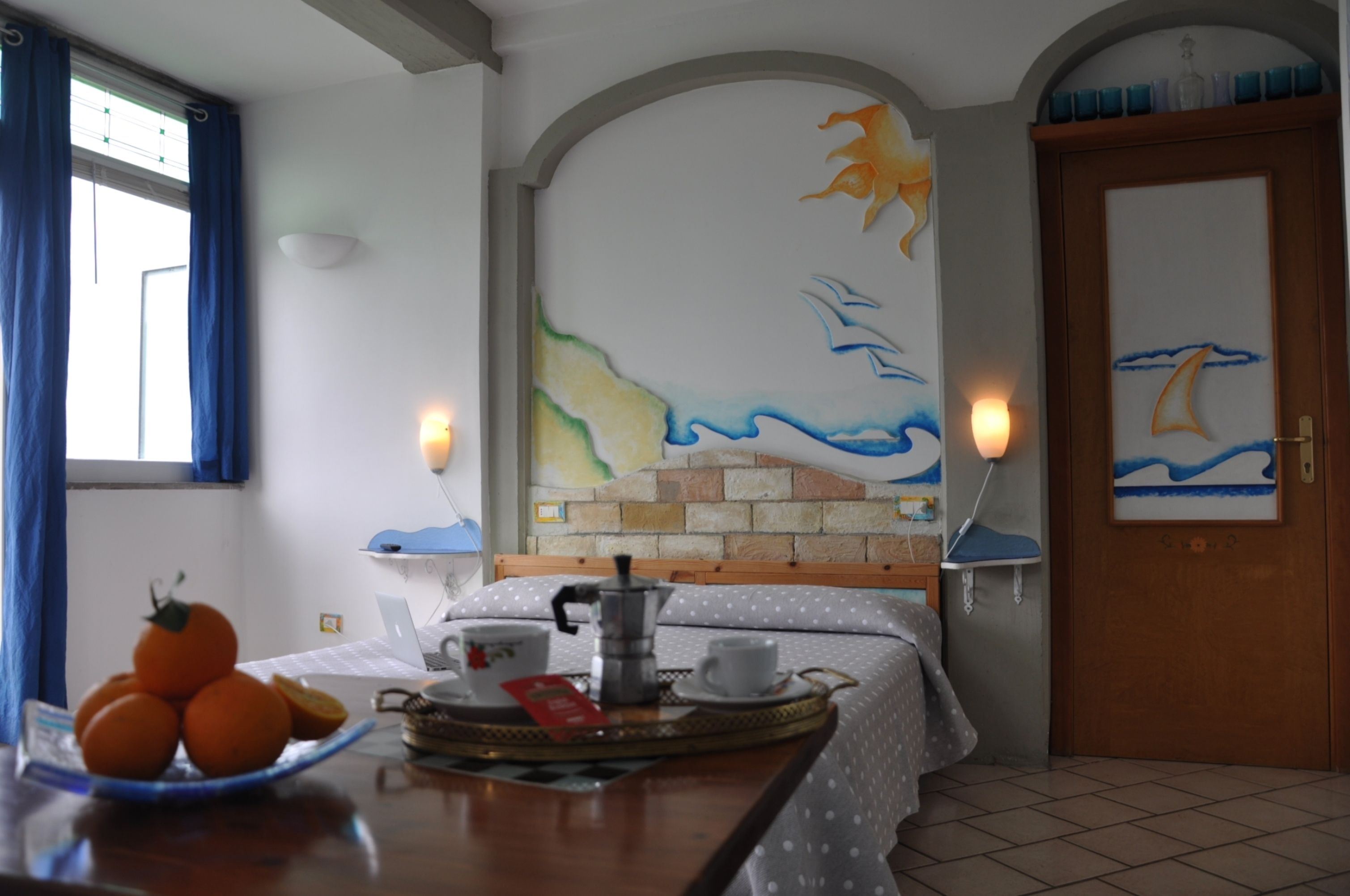A room with a bed, a table with oranges, a coffee maker, and a painting of a sailboat on the wall.