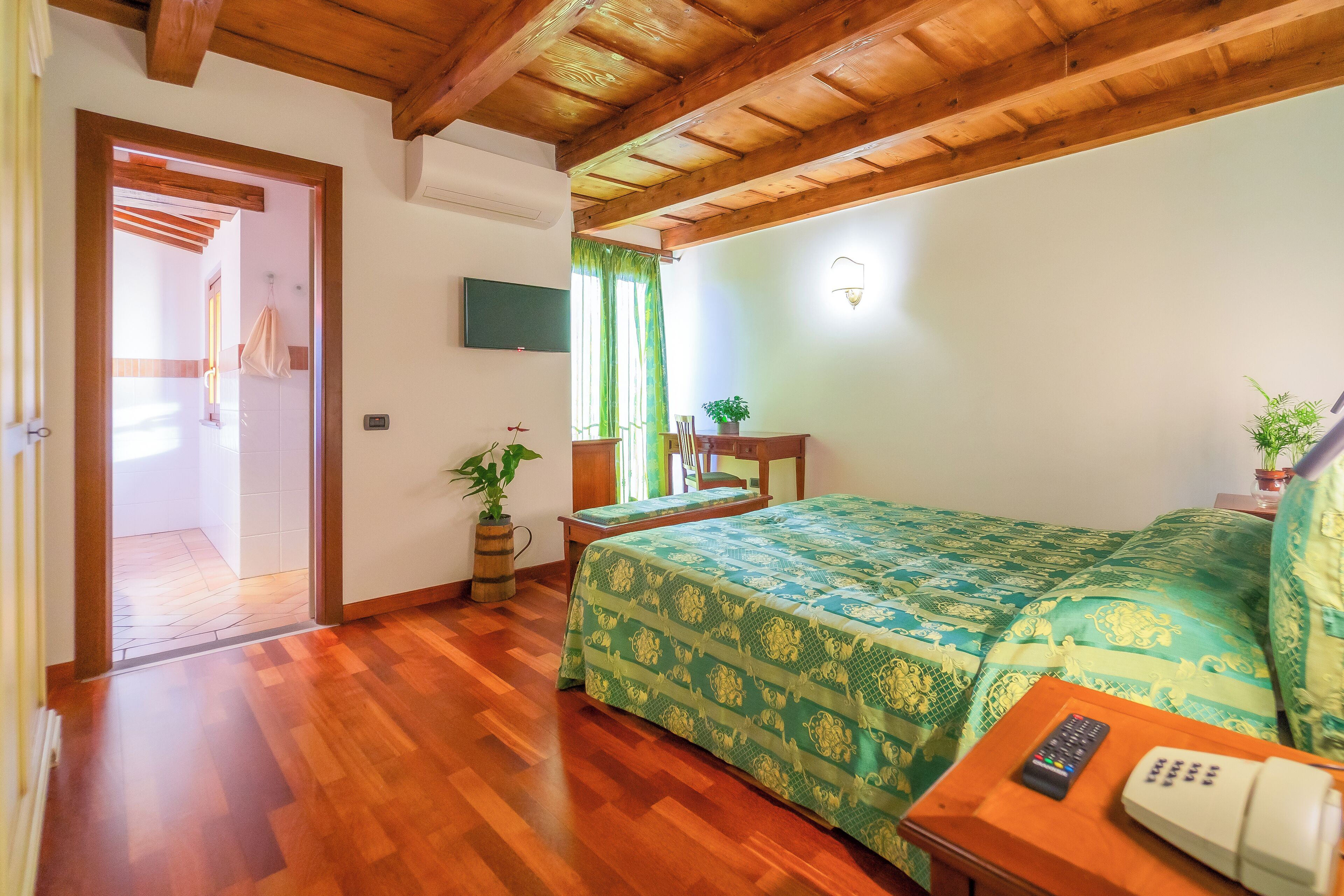 A room with wooden flooring, a bed with a green patterned bedspread, a bedside table with a remote control and a phone, a potted plant, and a television on the wall.