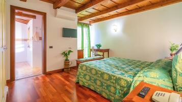 A room with wooden flooring, a bed with a green patterned bedspread, a bedside table with a remote control and a phone, a potted plant, and a television on the wall.