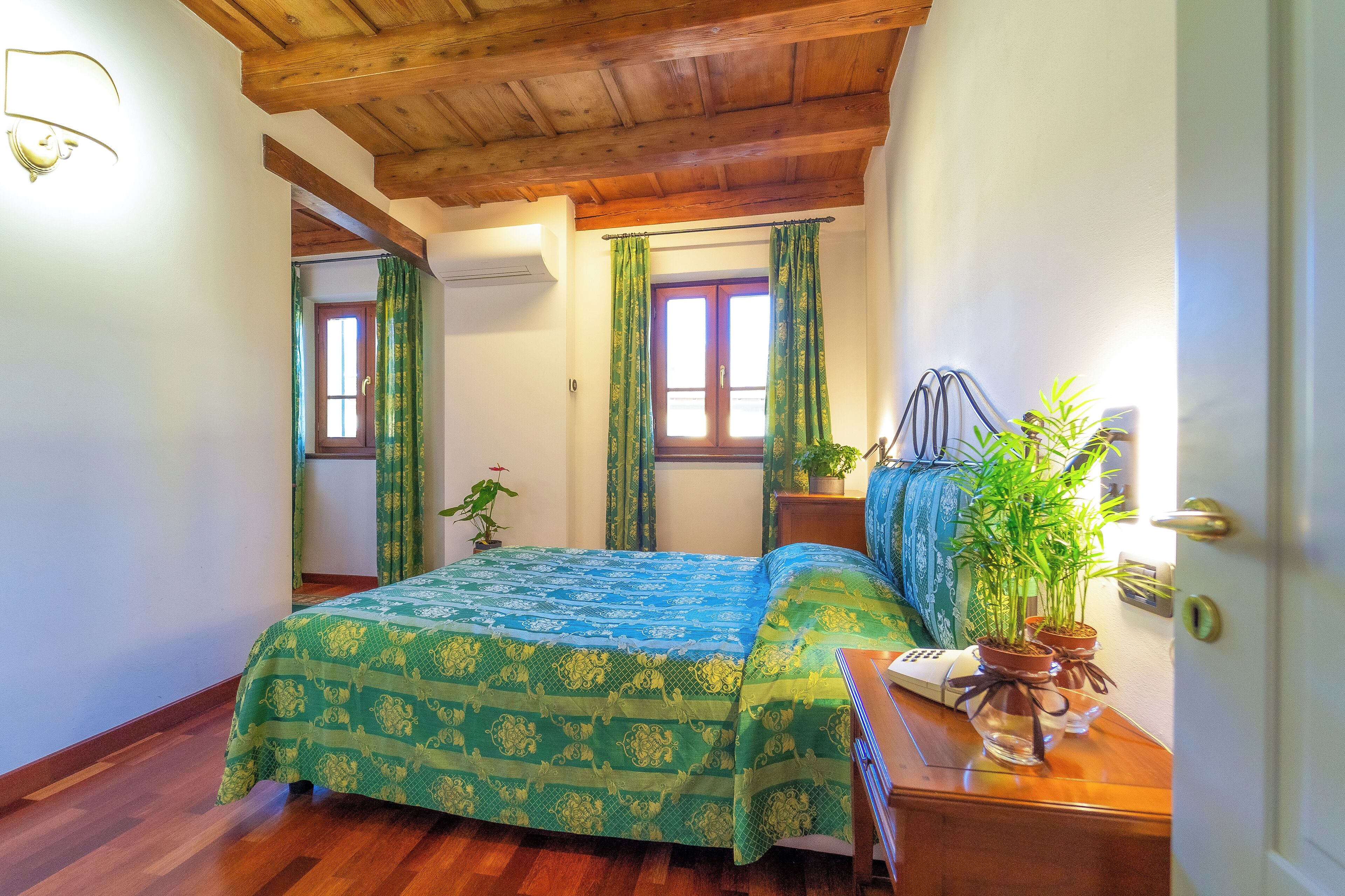 Ein Schlafzimmer mit Holzboden, einem Bett mit blaugrüner Bettwäsche, zwei Fenstern mit grünen Vorhängen und einem Nachttisch mit einer Pflanze.