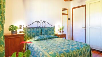 A bedroom with a wooden ceiling, a bed with patterned bedding, a bedside table with a lamp, and a potted plant.