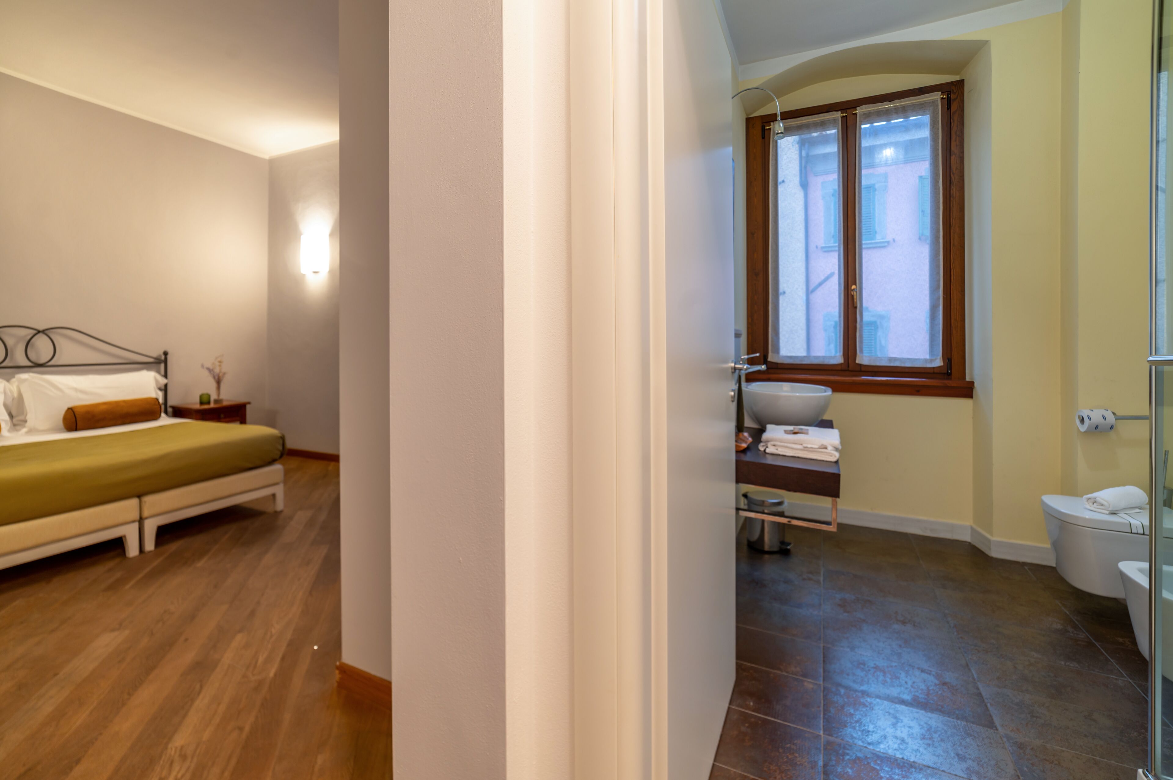Ein Schlafzimmer mit Bett, Nachttisch und Ablagefläche. Ein Badezimmer ist sichtbar, mit Waschbecken, Spiegel und Toilette.